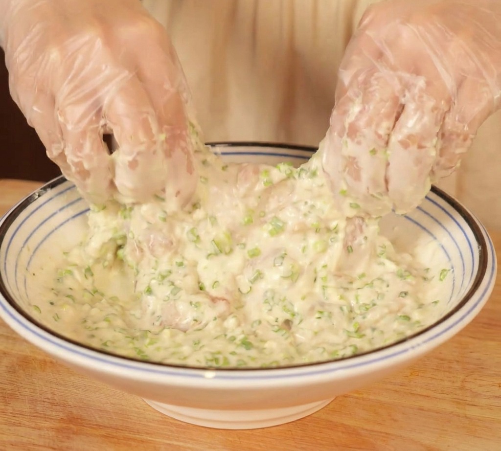 Hände in Handschuhen wenden die Hähnchenstücke in einem dicken weißen Teig voller grüner Frühlingszwiebelstücke.