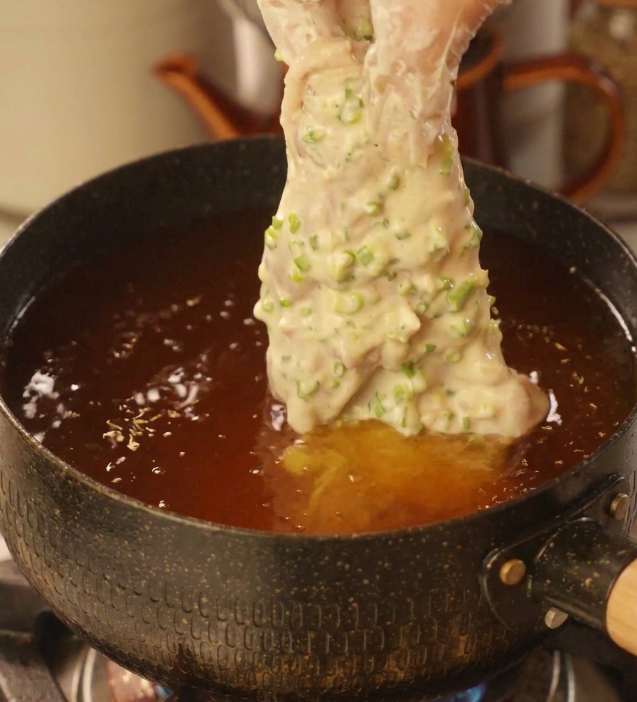 Eine Hand lässt ein dick mit Frühlingszwiebel-Teig ummanteltes Hähnchenstück vorsichtig in einen Topf mit heißem Öl gleiten.