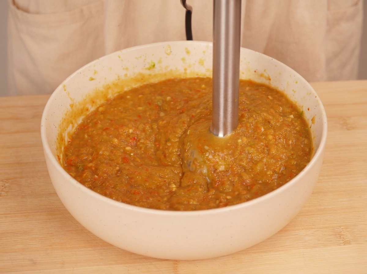 Ein Pürierstab verarbeitet geröstetes Gemüse in einer weißen Schüssel zu einer dickflüssigen, orange-grünen Sauce.