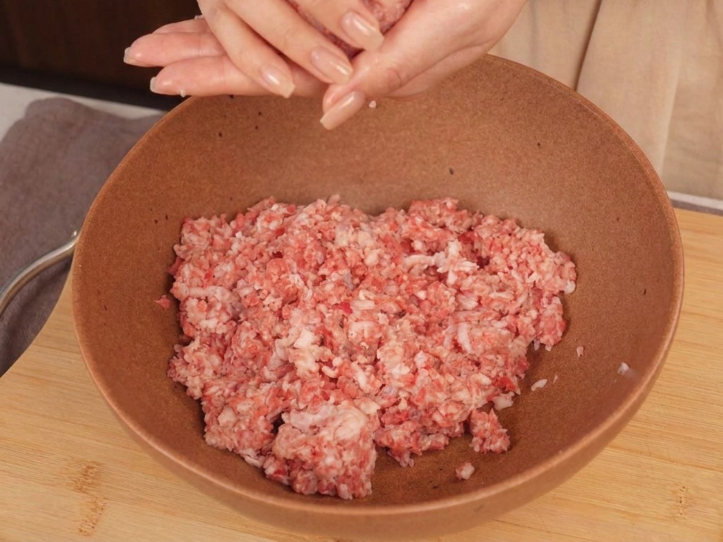 Hände formen aus rohem Rinderhackfleisch eine runde Kugel über einer Schüssel.