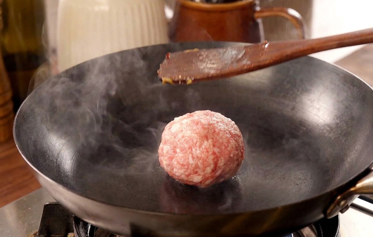 Ein Pfannenwender drückt ein Fleischbällchen in einer heißen Pfanne flach, während Dampf aufsteigt.
