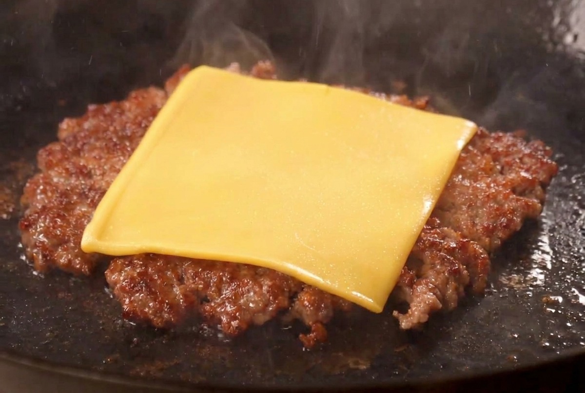 Eine Scheibe gelber Schmelzkäse schmilzt auf einem kross gebratenen Burger-Patty in der Pfanne.
