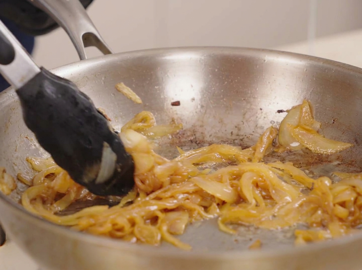 Eine Grillzange rührt in einer dichten, glänzenden Menge tiefgoldener karamellisierter Zwiebeln in einer Pfanne.