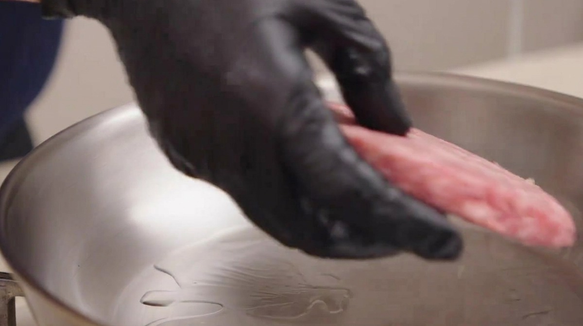 Eine Hand mit schwarzem Küchenhandschuh legt vorsichtig ein rohes, marmoriertes Rinder-Patty in eine glänzende Pfanne mit Öl.