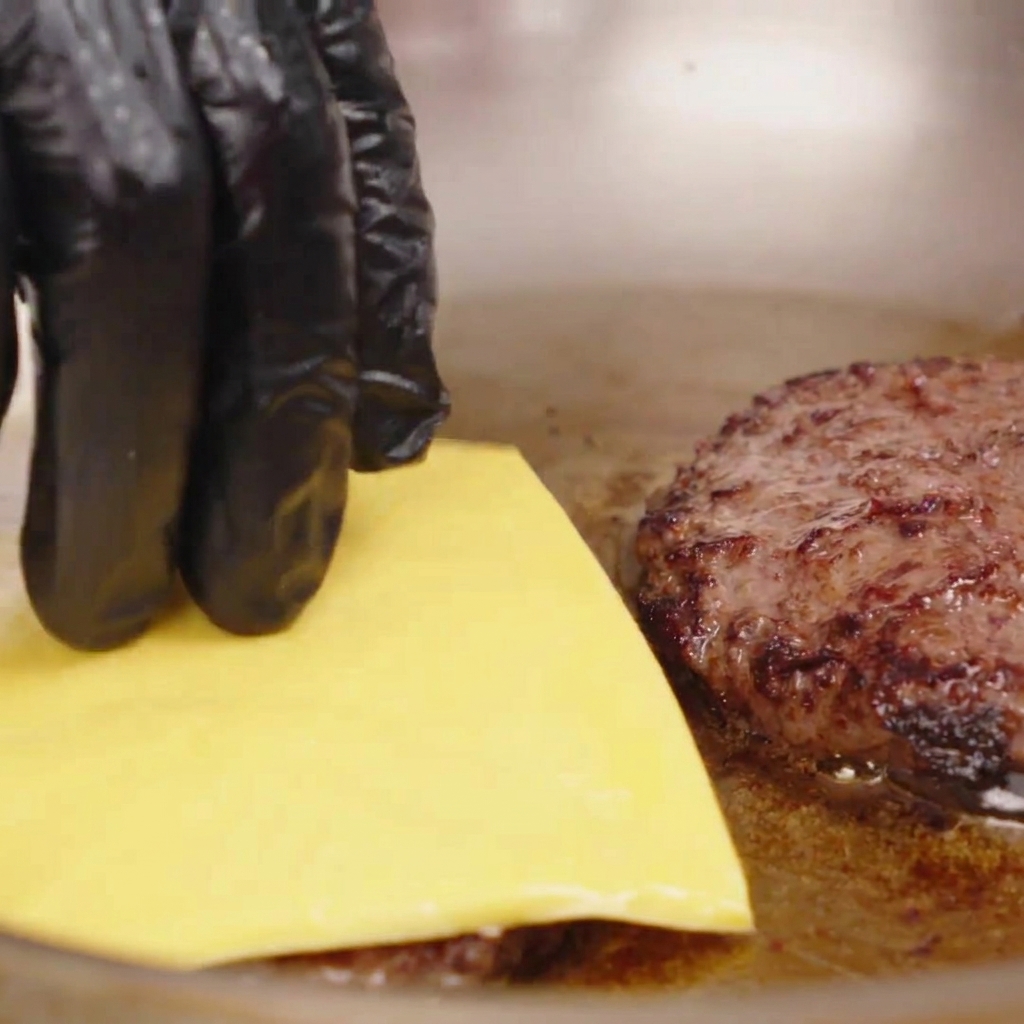 Eine Hand legt eine quadratische Scheibe gelben Käse auf ein heißes, angebratenes Rindfleisch-Patty in einer Pfanne.