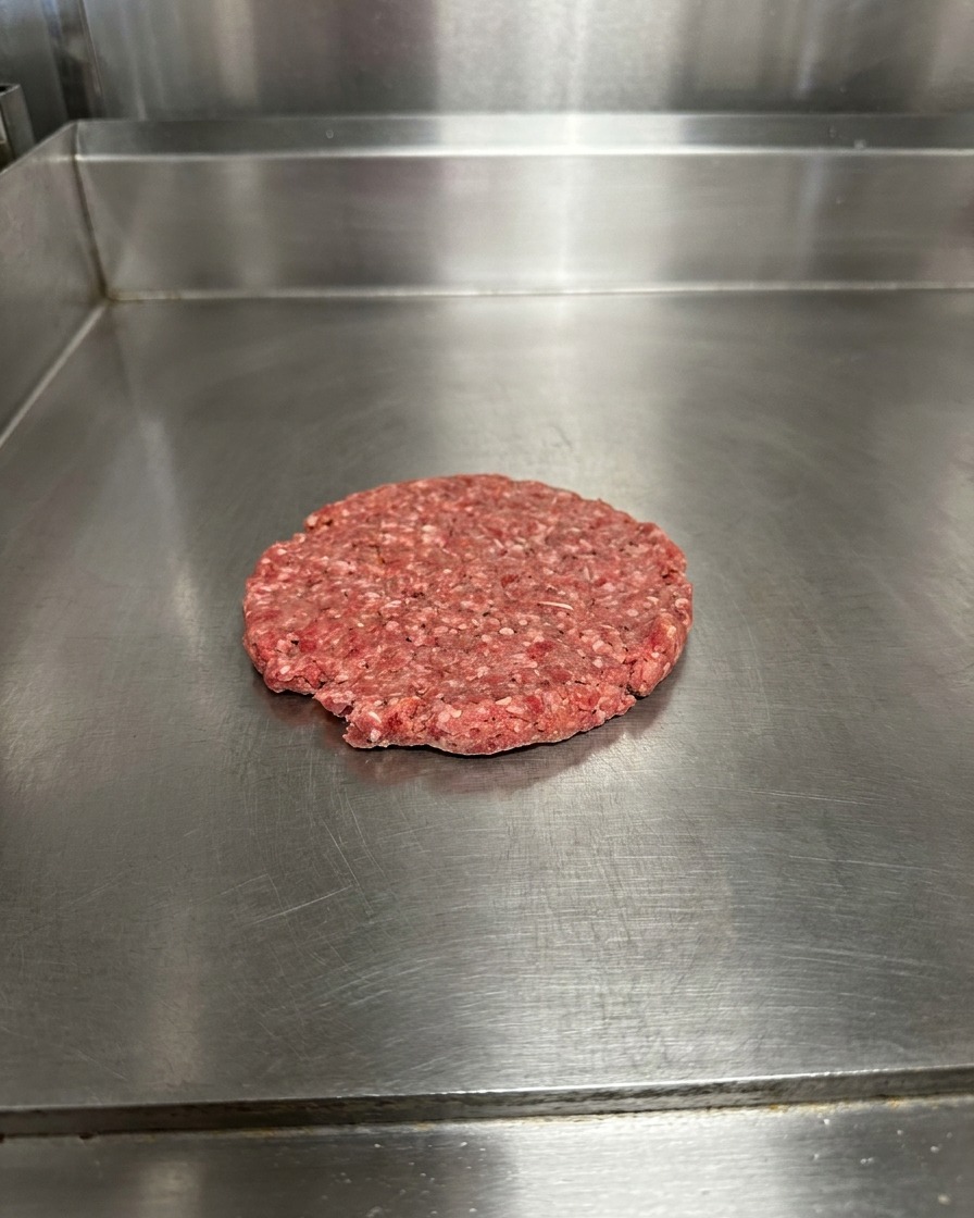 Ein flach gedrücktes, rohes Patty auf einer Metalloberfläche, bestreut mit Salz und Pfeffer.