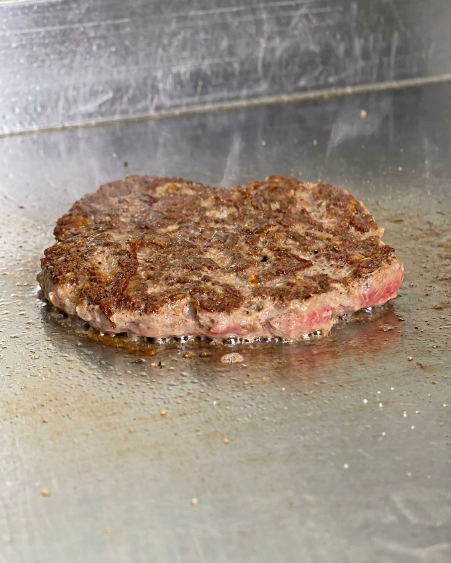 Ein gewendetes Burger-Patty auf dem Grill mit einer dunkelbraunen, knusprigen Kruste und brutzelndem Fett.