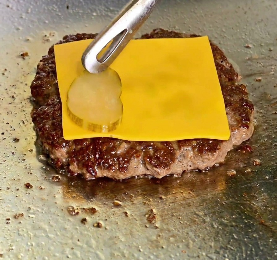 Eine Scheibe geschmolzener gelber Cheddar auf einem Patty, während eine Gurkenscheibe darauf platziert wird.