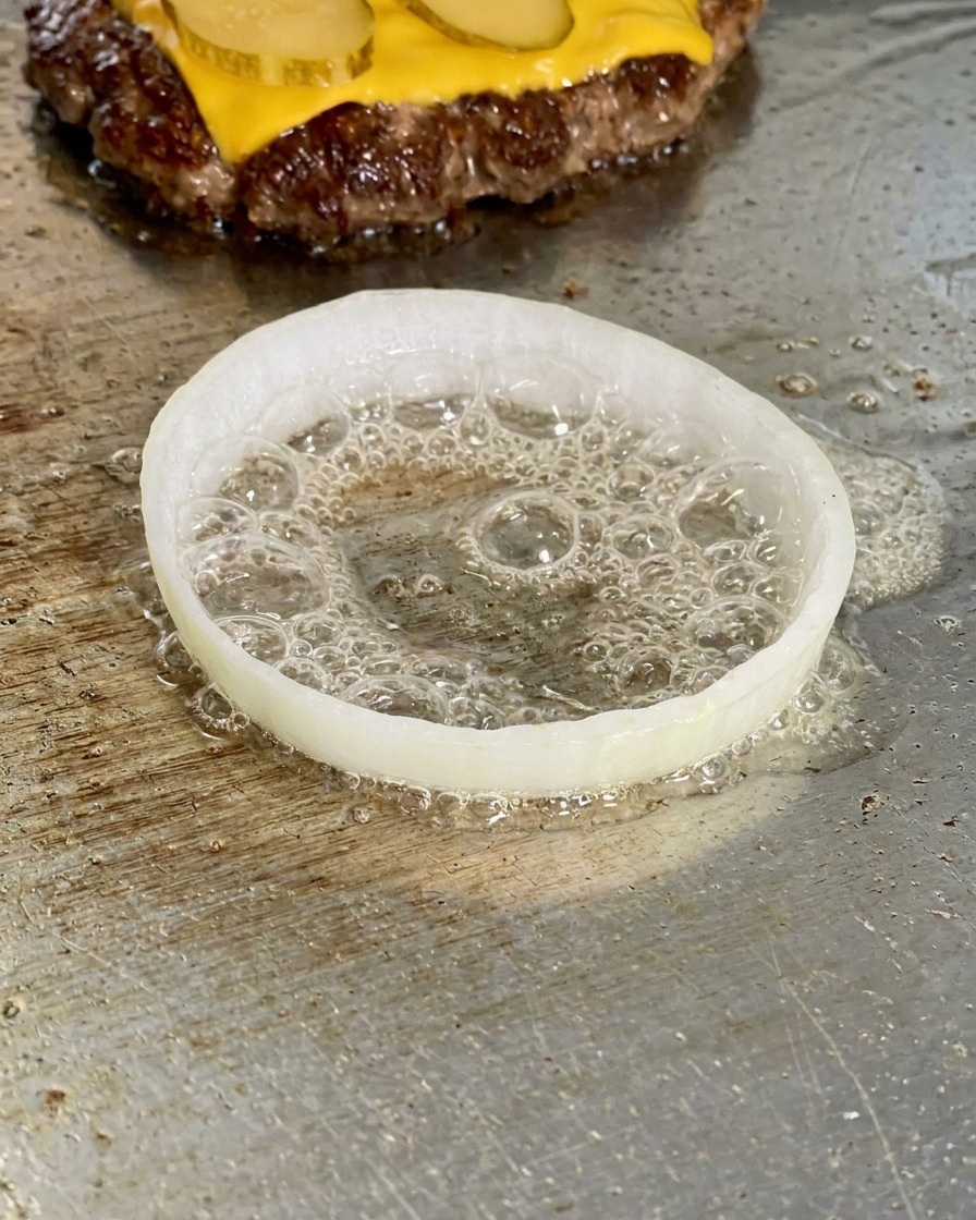 Ein Zwiebelring auf der Grillplatte, in dessen Mitte ein Ei in sprudelndem Öl brät.