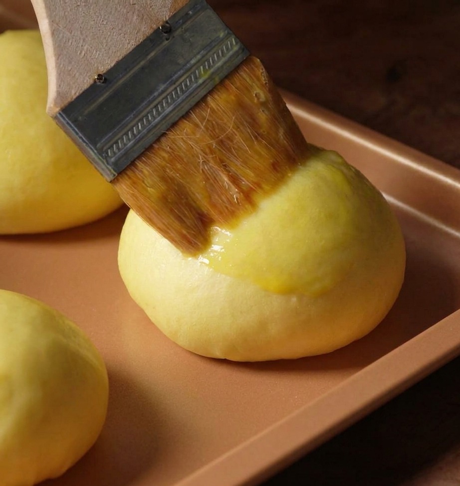 Ein Backpinsel trägt eine gelbe Eigelb-Mischung auf einen runden Brioche-Teigling auf.