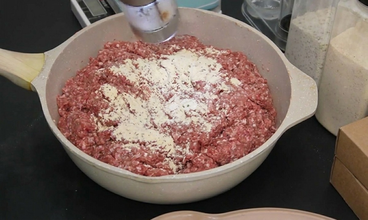 Eine Person mit schwarzem Handschuh verwendet eine elektrische Mühle, um Meersalz zum rohen Rinderhackfleisch hinzuzufügen.