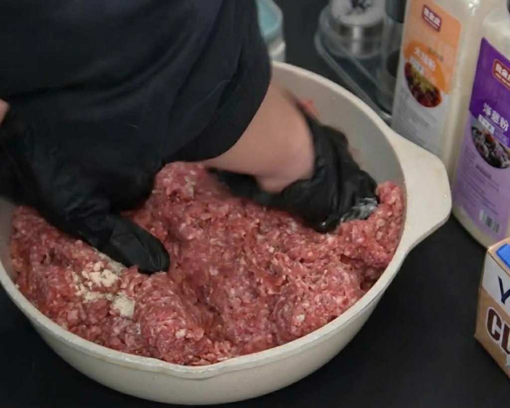 Hände in schwarzen Handschuhen mischen und kneten das gewürzte rohe Rinderhackfleisch in einer hellen Pfanne.