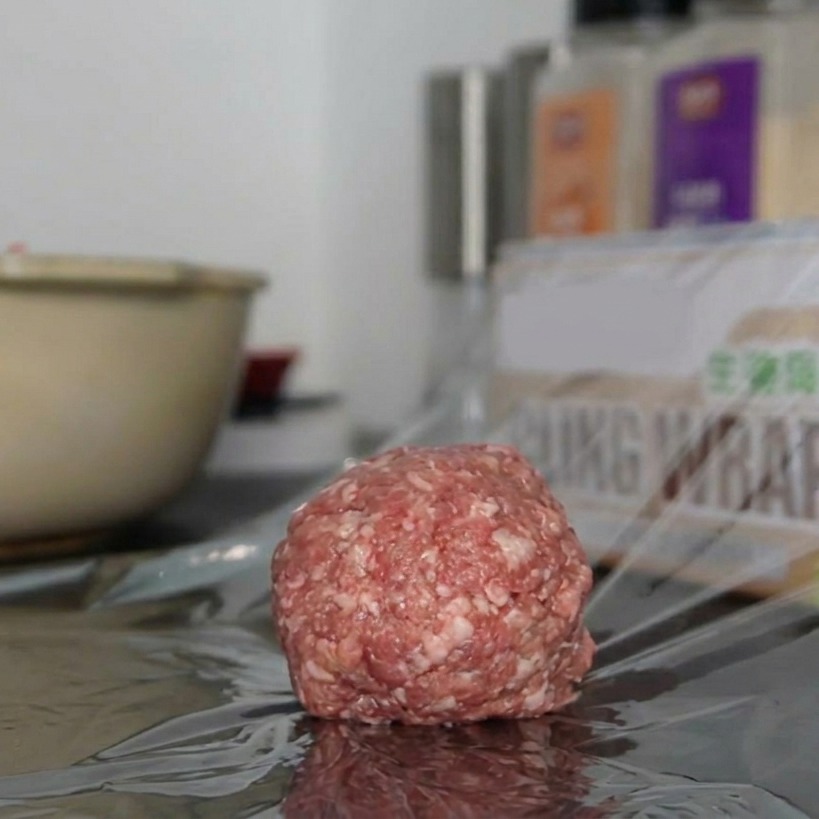 Eine einzelne, runde Portion gewürztes Rinderhackfleisch ruht auf einem Stück transparenter Frischhaltefolie auf einer Arbeitsplatte.