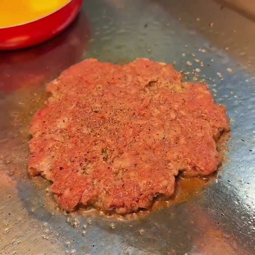 Ein Rindfleisch-Patty brät auf einem flachen Grill und wird mit Salz und Pfeffer gewürzt.