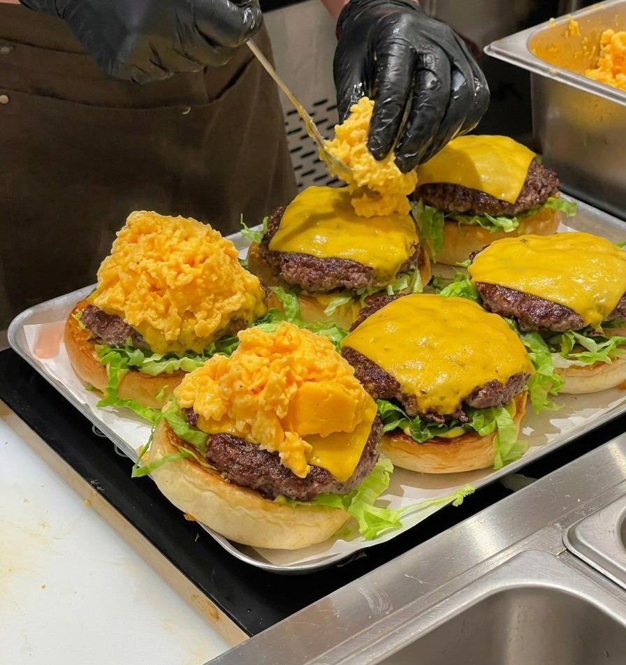 Ein Koch gibt mit einem Löffel eine große Portion Rührei auf die Cheeseburger.
