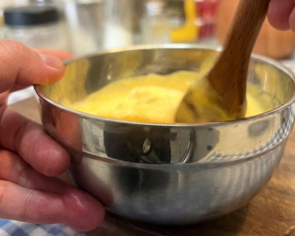 Eine Hand rührt mit einem Holzlöffel eine cremige, orange-gelbe Burgersoße in einer kleinen Edelstahlschüssel um.