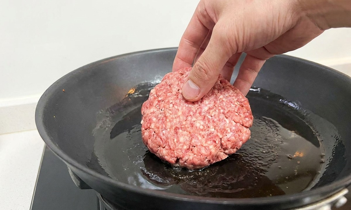 Eine Hand legt ein rohes Rindfleisch-Patty in eine heiße, schwarze Pfanne mit schimmerndem Öl.