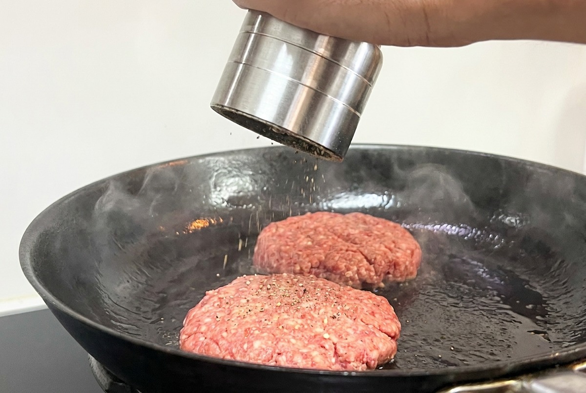 Eine Hand würzt zwei bratende Rindfleisch-Patties in der Pfanne mit einer Pfeffermühle.