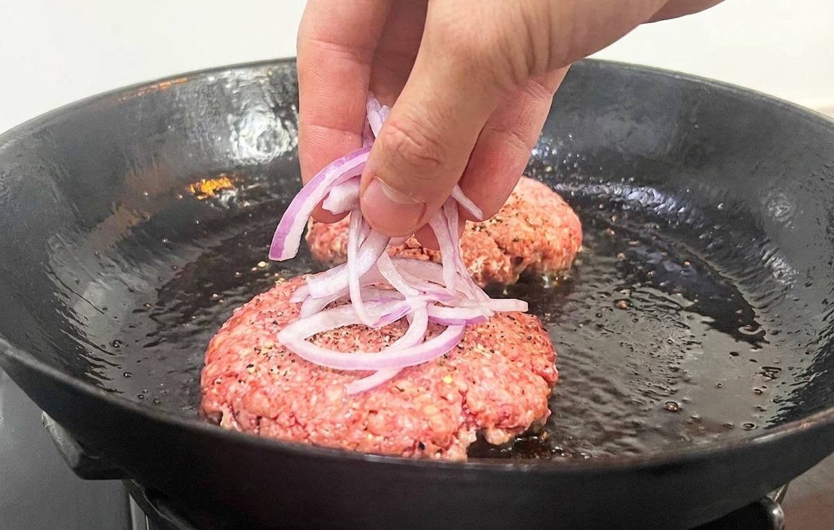 Eine Hand verteilt feine Ringe einer roten Zwiebel auf den rohen Rindfleisch-Patties in einer Bratpfanne.