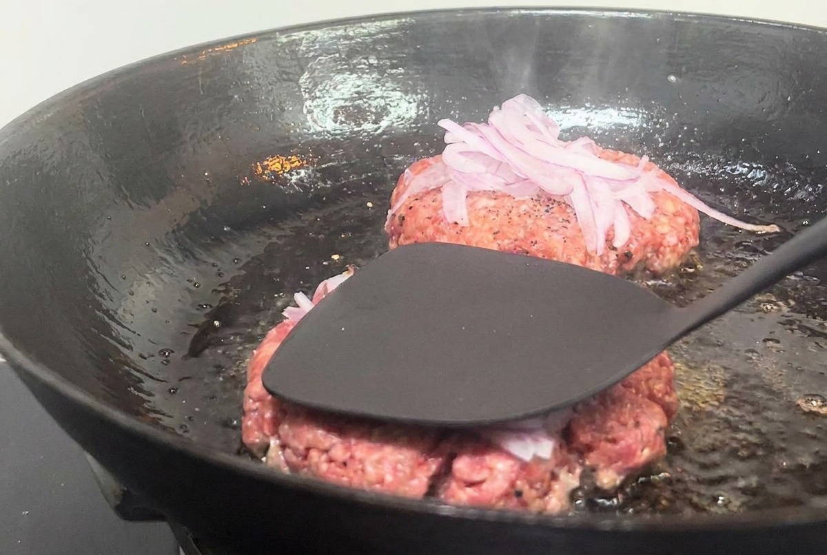 Ein schwarzer Metallspatel drückt ein mit Zwiebeln belegtes Burger-Patty in einer heißen Pfanne platt.