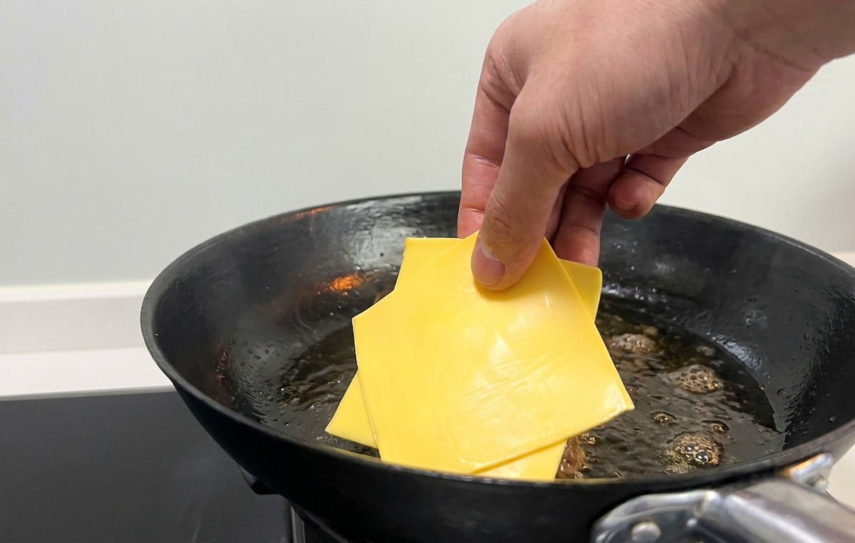 Eine Hand legt quadratische Scheiben gelben Käse auf die gebratenen Burger-Patties in der Pfanne.