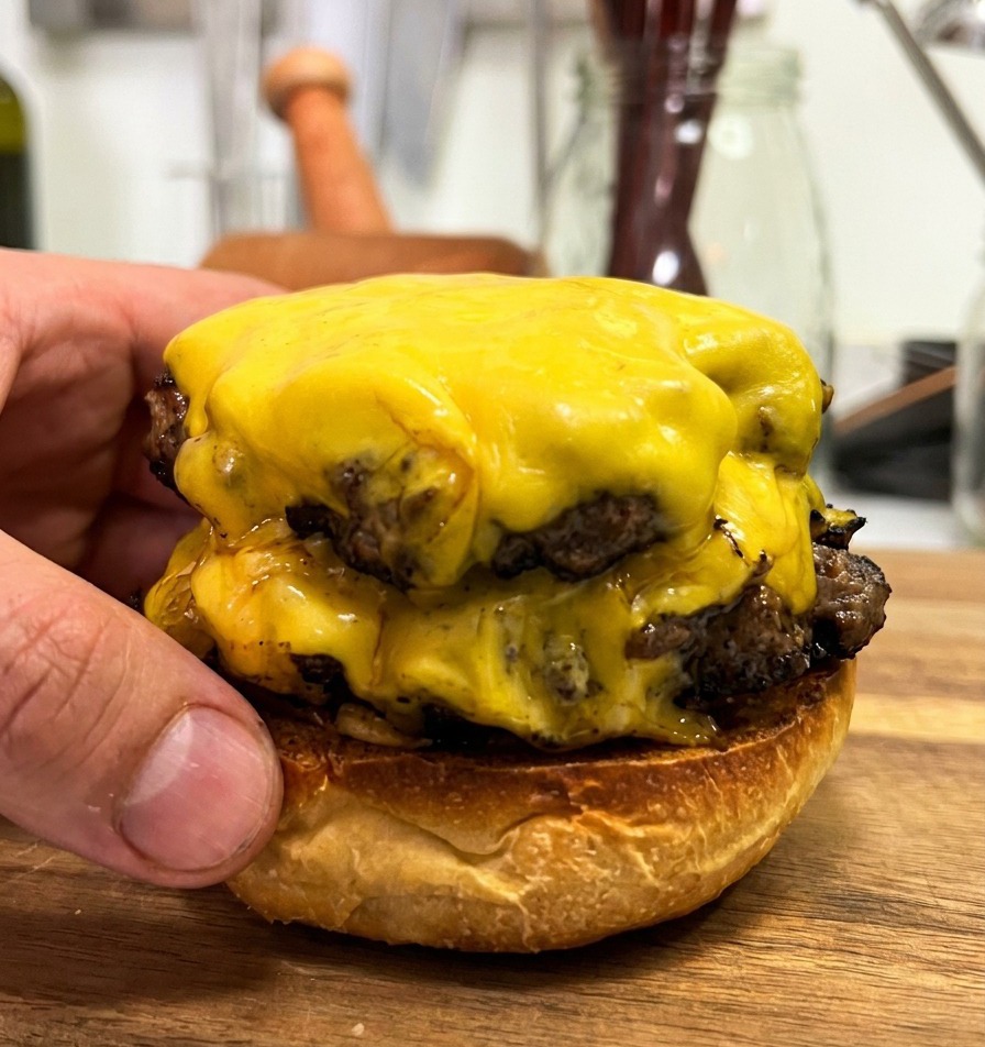 Zwei dicke Rindfleisch-Patties mit zerlaufenem Käse werden auf dem unteren Bun platziert.