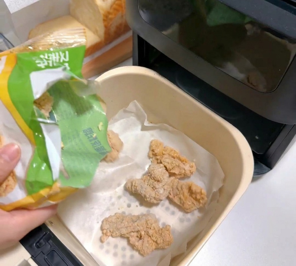 Tiefgekühlte panierte Hähnchenteile werden in einen mit Backpapier ausgelegten Korb der Heißluftfritteuse geschüttet.