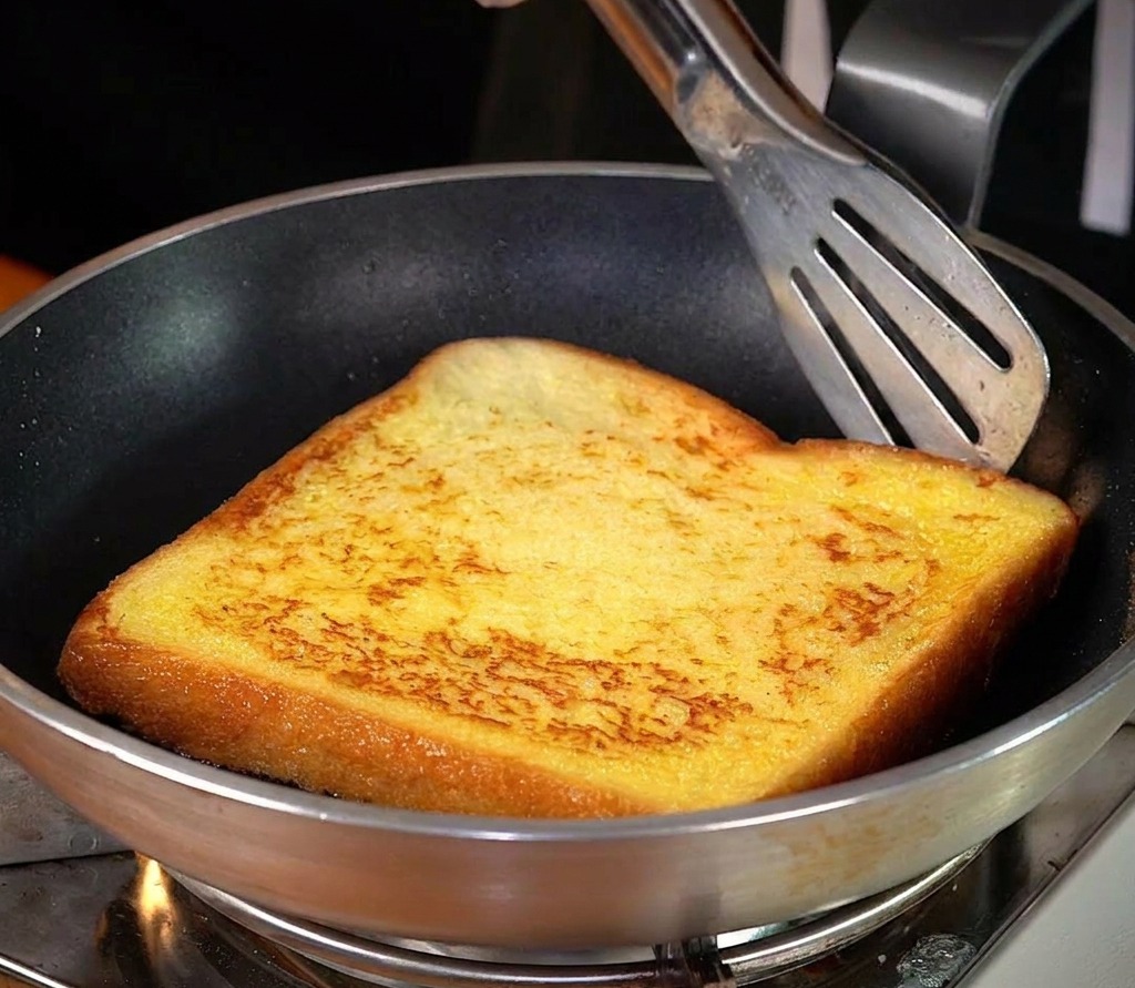 Eine einzelne Scheibe dicker Toast wird in einer silbernen Pfanne in geschmolzener Butter gebraten und zeigt eine goldbraune Kruste.