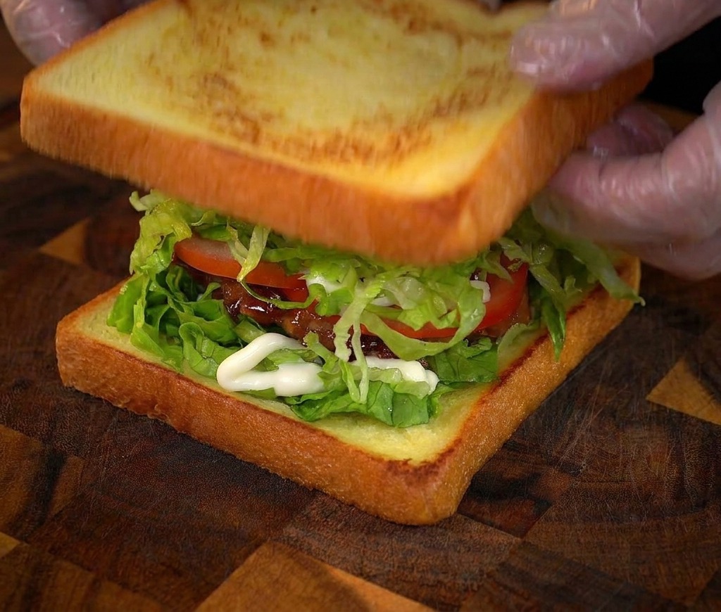 Hände legen die letzte Scheibe geröstetes Brot auf ein hohes, mehrschichtiges Hähnchen-Sandwich.