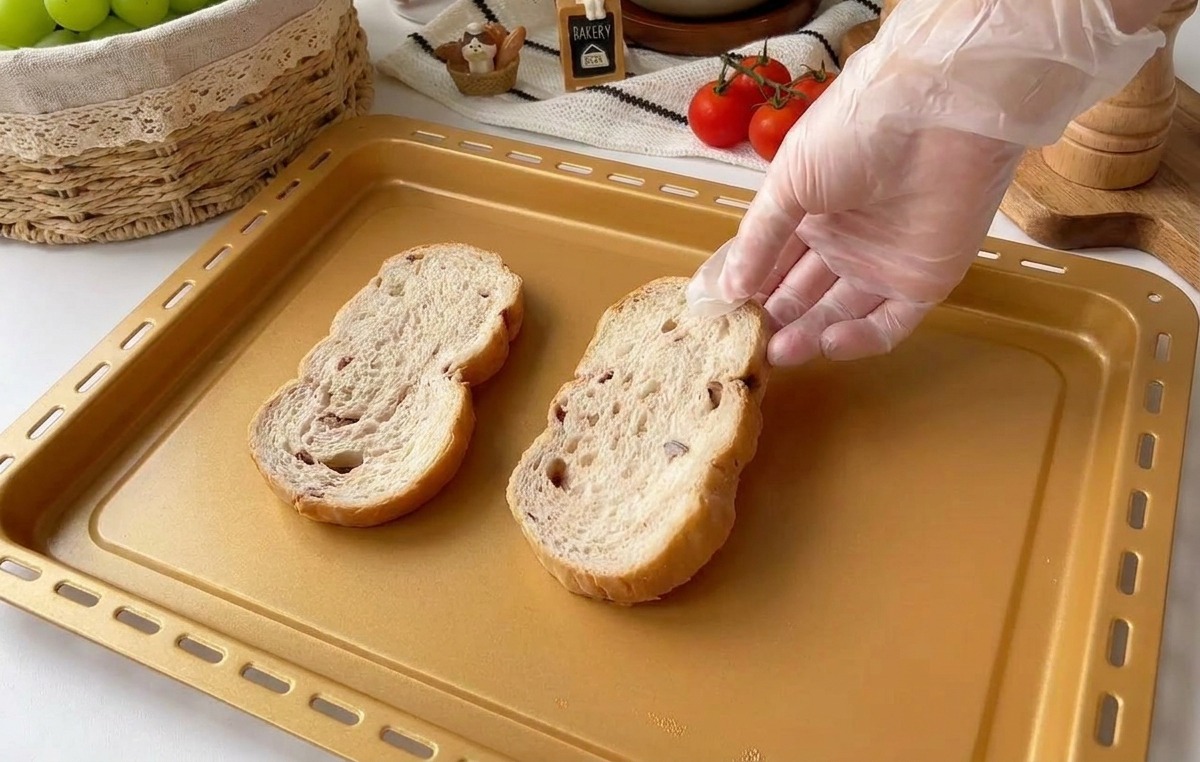 Eine Hand in einem Klarsichthandschuh legt zwei dicke Scheiben rustikales Mehrkornbrot auf ein goldfarbenes Backblech.