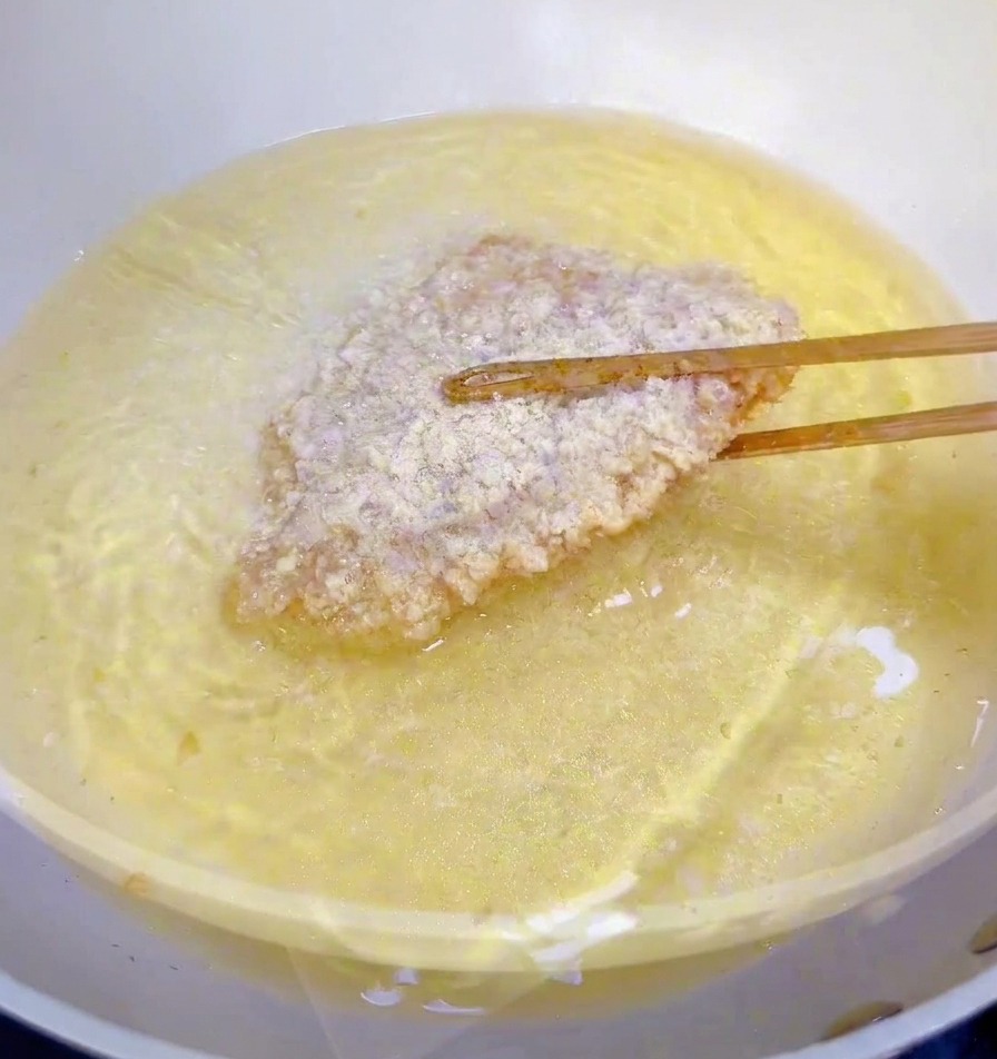 Ein paniertes Hähnchenfilet wird mit Stäbchen in eine Pfanne mit heißem Öl gleiten gelassen.