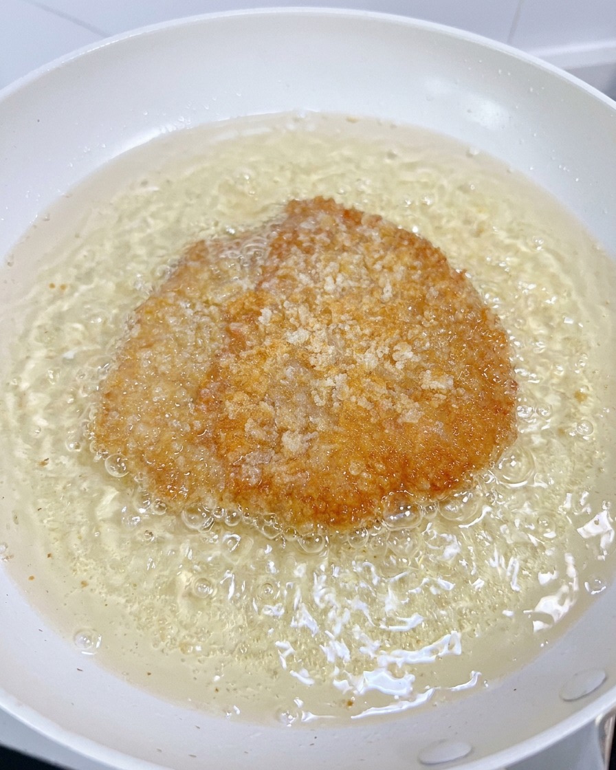 Ein Hähnchenfilet brutzelt in einer weißen Pfanne, während die Panade goldgelb wird.