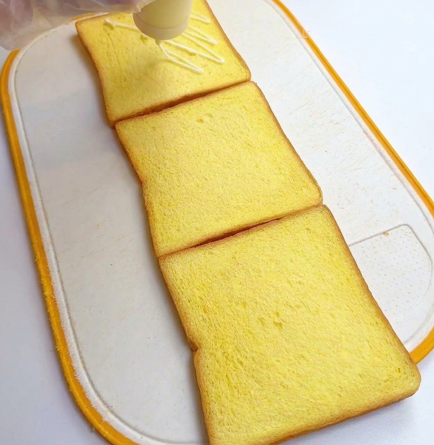 Eine Person verteilt weißes Dressing aus einer Flasche auf drei quadratischen Toastscheiben.