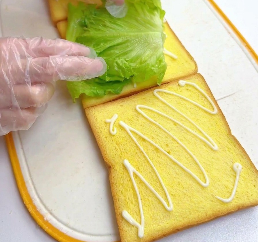 Hände legen ein grünes Salatblatt auf eine Toastscheibe mit Dressing.