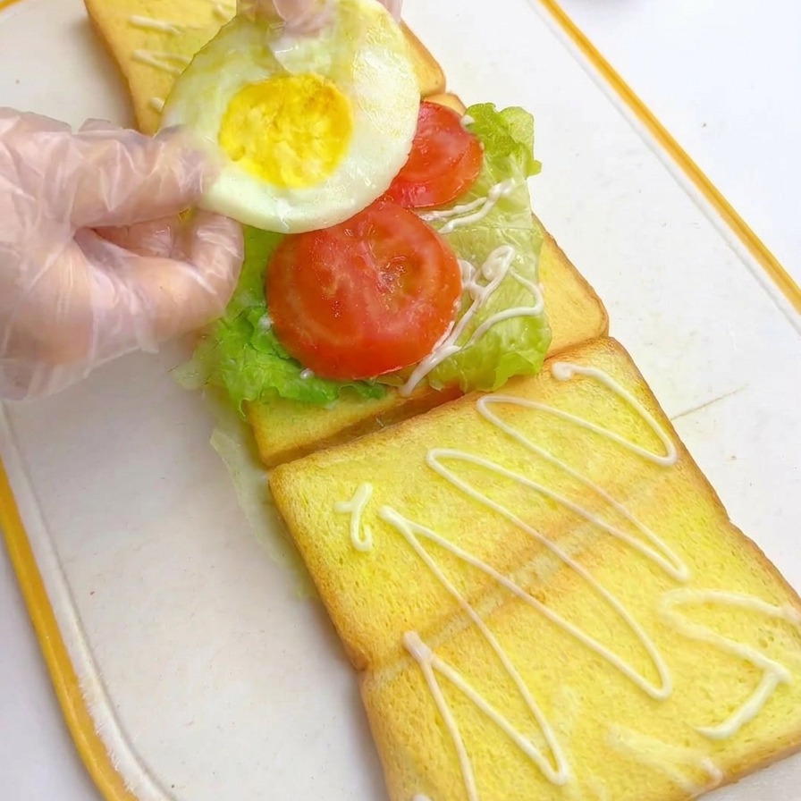 Hände platzieren ein gebratenes Ei auf die Tomatenscheiben in der Sandwich-Schichtung.