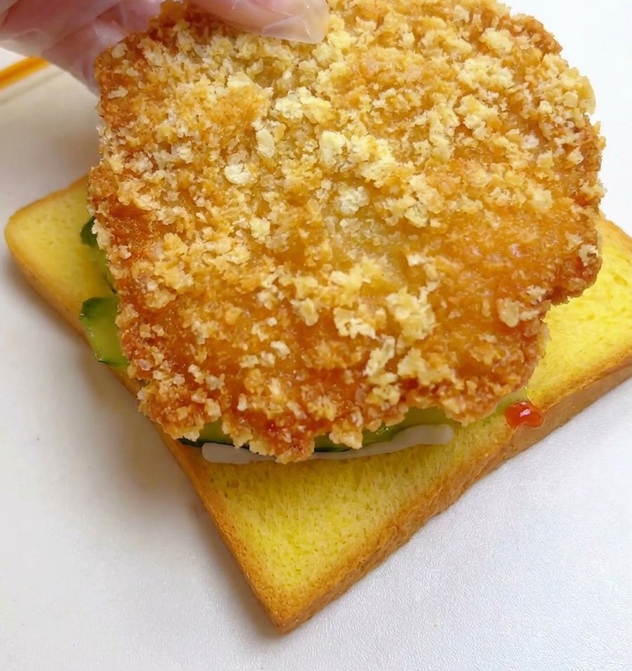 Eine Hand legt das goldbraun frittierte Hähnchenfilet auf die Gurkenscheiben.