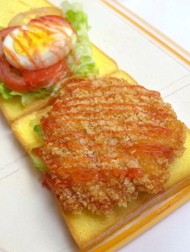 Ketchup wird über das Hähnchenfilet auf dem Toast gegeben, im Hintergrund sind weitere Schichten zu sehen.