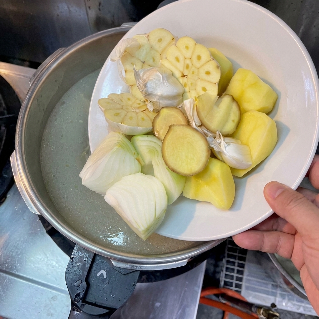 Eine Hand schiebt große Kartoffelstücke, halbierte Zwiebeln, Ingwer und halbierte Knoblauchknollen von einem Teller in einen Topf mit trüber Brühe.