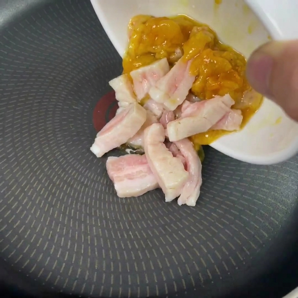Eine Hand gibt rohe Stücke von weißem und gelbem Fett aus einer kleinen Schale in eine kalte Bratpfanne.
