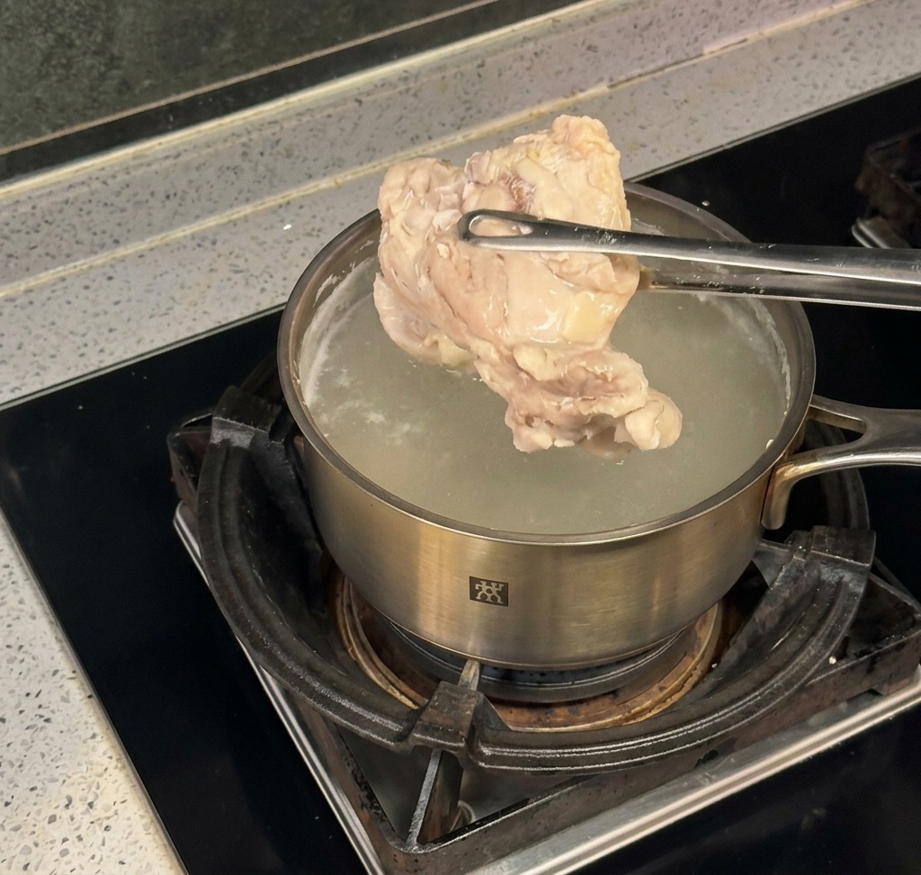 Eine Metallzange hebt ein großes Stück gekochtes Hähnchen aus einem Edelstahltopf mit kochendem Wasser auf einem Herd.