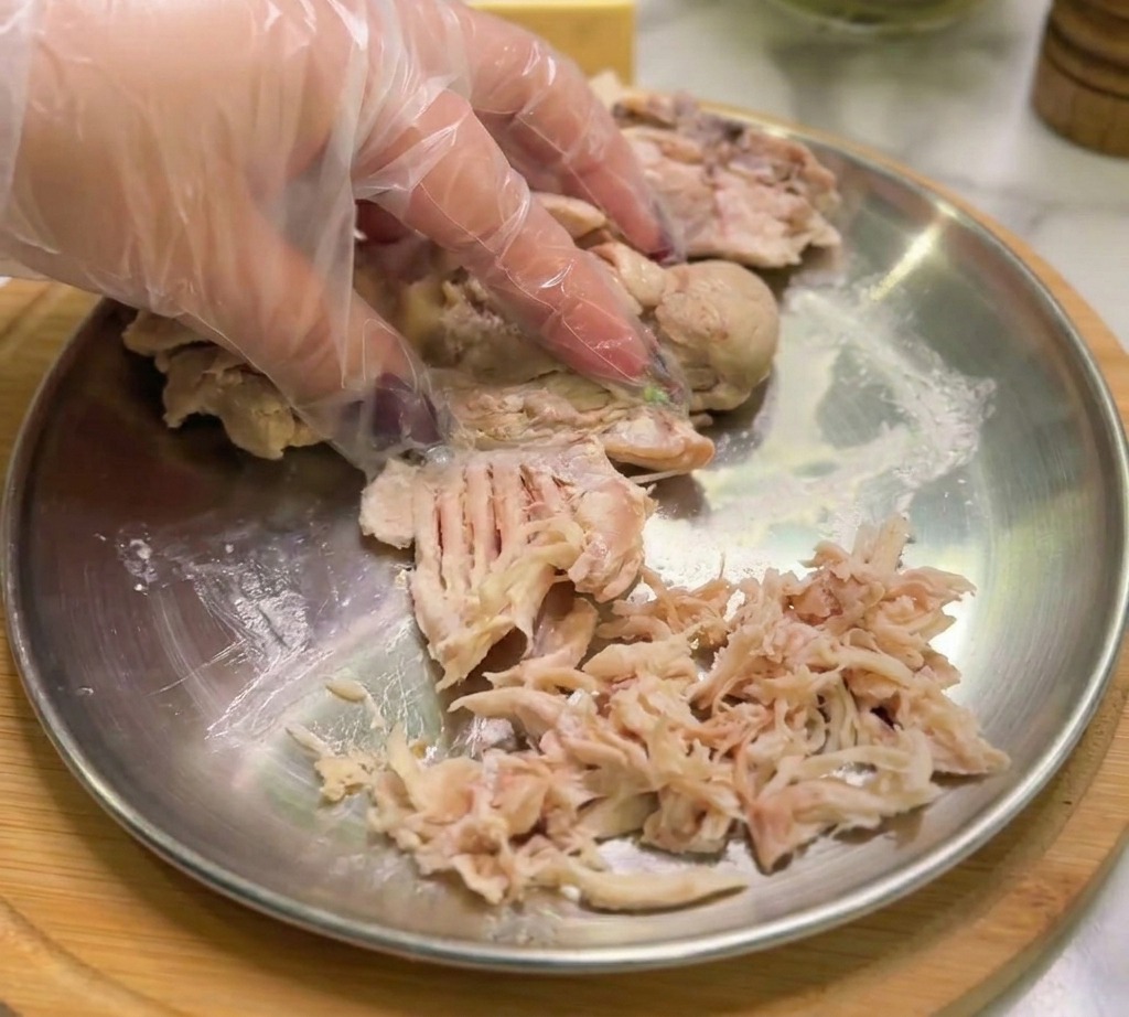 Eine Hand mit Handschuh zupft gekochtes Hähnchenfleisch in feine Streifen auf einer flachen Metallplatte.