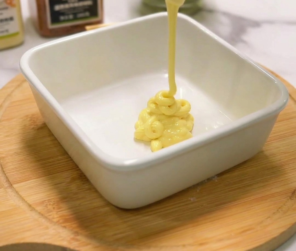 Cremige gelbe Mayonnaise wird aus einer Flasche in eine kleine weiße Keramikschale auf einem Holzbrett gedrückt.