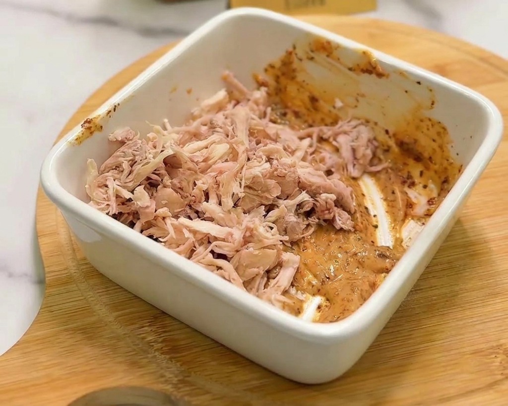 Ein Haufen zerkleinertes Hähnchenfleisch wird in einer weißen Keramikschale mit der orangefarbenen Würzsauce vermischt.