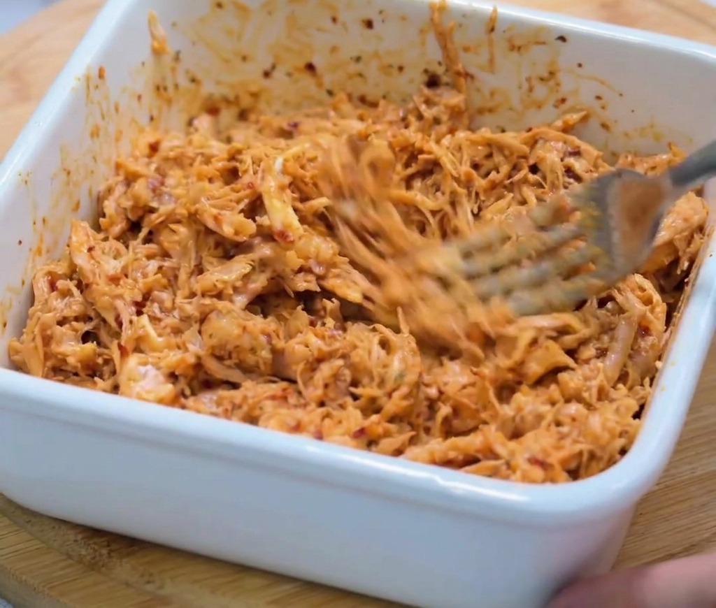 Nahaufnahme: Fein zerkleinertes Hähnchen wird mit einer Metallgabel kräftig in einer dicken, roten Würzsauce verrührt.
