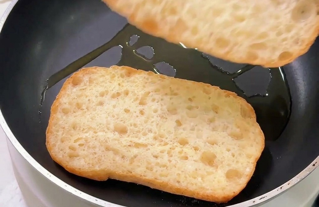 Zwei Stücke Ciabatta-Brot werden mit der Schnittseite nach unten in einer Pfanne mit etwas Öl geröstet.