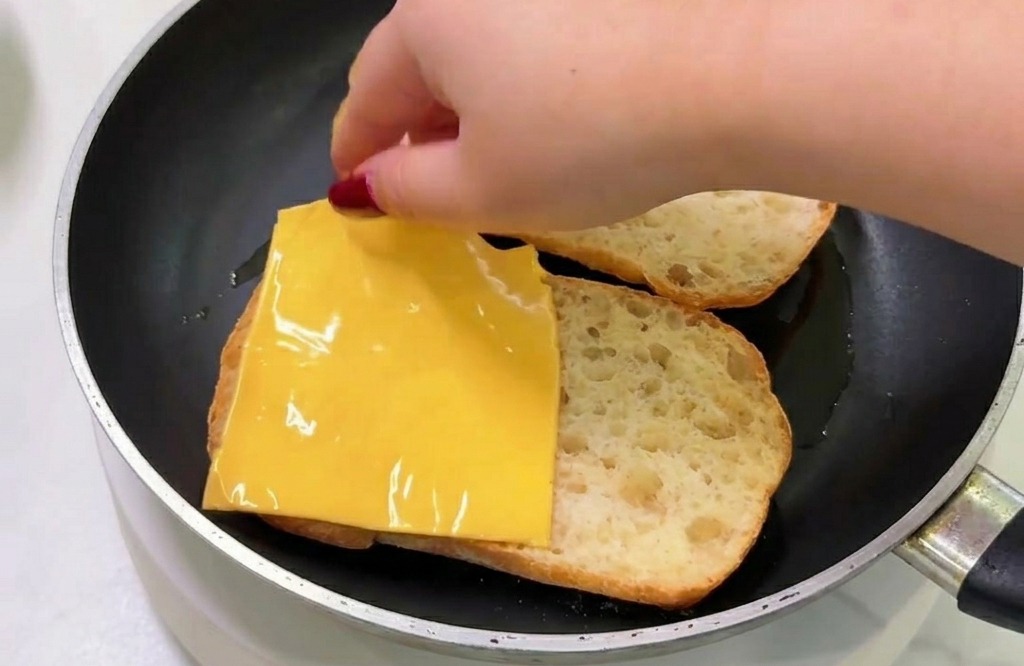 Eine Hand legt eine quadratische Scheibe gelben Schmelzkäse auf ein Stück geröstetes Brot in einer Bratpfanne.