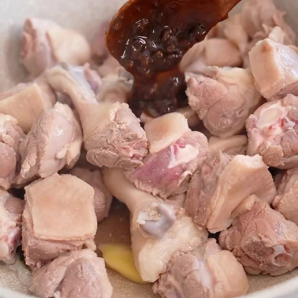 Ein Holzlöffel gibt dunkle Bohnenpaste über Entenstücke, die in einer hellen Pfanne gebraten werden.