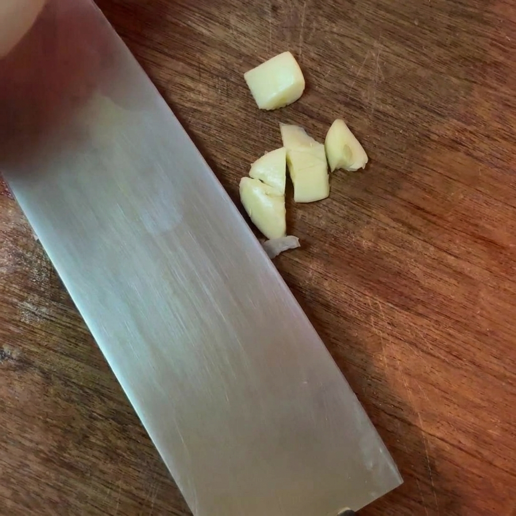 Grob gehackte Knoblauchzehen neben einem großen Metallmesser auf einem Holzschneidebrett.