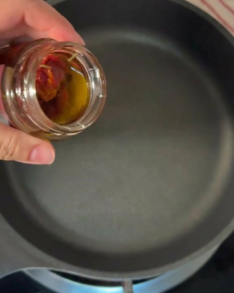 Eine Hand neigt ein kleines Glas, um orange getöntes Öl in eine erhitzte schwarze Pfanne zu gießen.