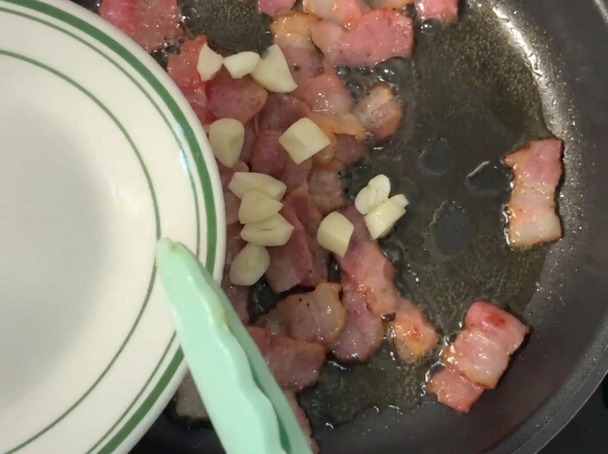 Frisch gehackter Knoblauch wird von einem Teller in eine Pfanne mit brutzelndem Speck geschoben.